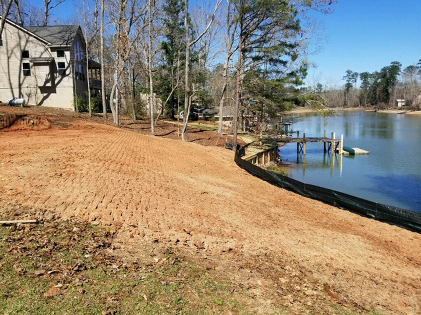 Modern lakeside house with light siding, large windows, surrounded by tall trees, grassy yard sloping toward water, wooden dock extending into lake