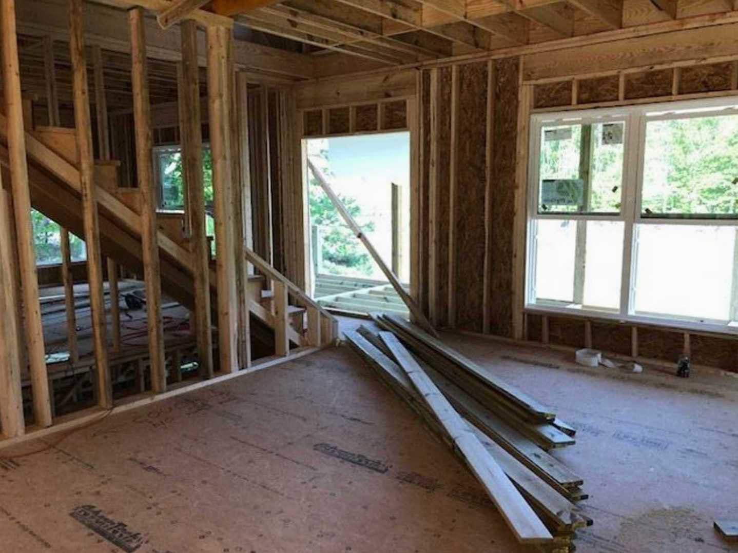 Open room featuring exposed wood ceiling beams, hardwood floors, wooden staircase, stacked lumber planks on the floor, multiple windows with metal bars, and construction materials