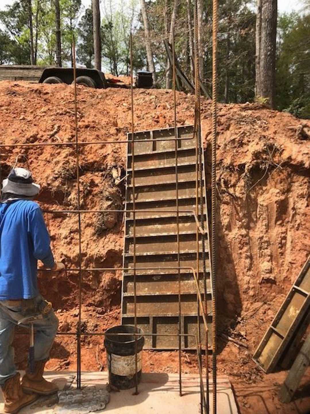 Stone exterior wall with rusted metal stairs and railing, man in work boots and blue jeans standing beside ladder, outdoor setting with trees and ground visible