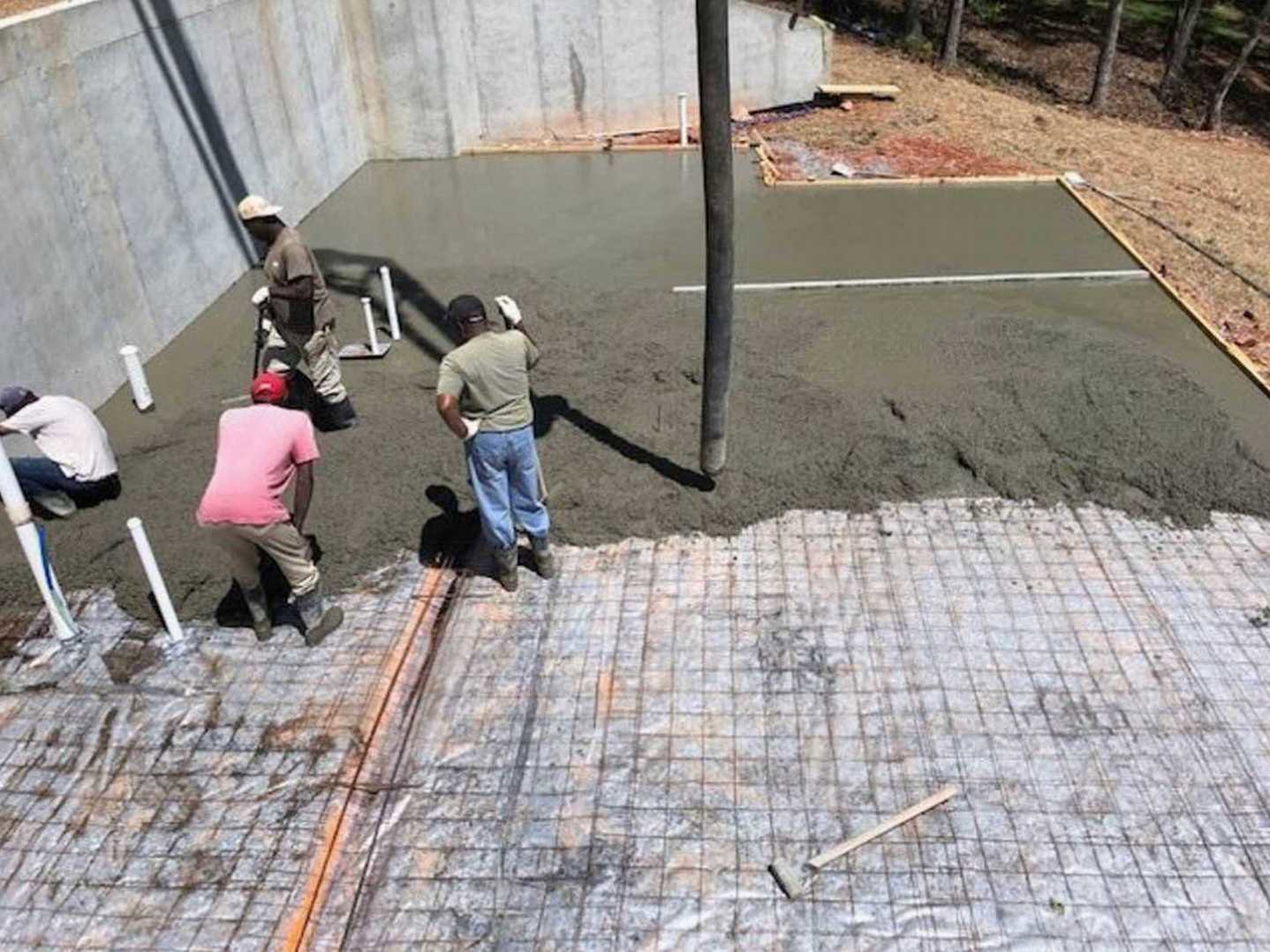 Crew pouring and smoothing concrete slab foundation outdoors, men in work clothes and boots, one in pink shirt, another bending down in white shirt, tarp and scooter nearby