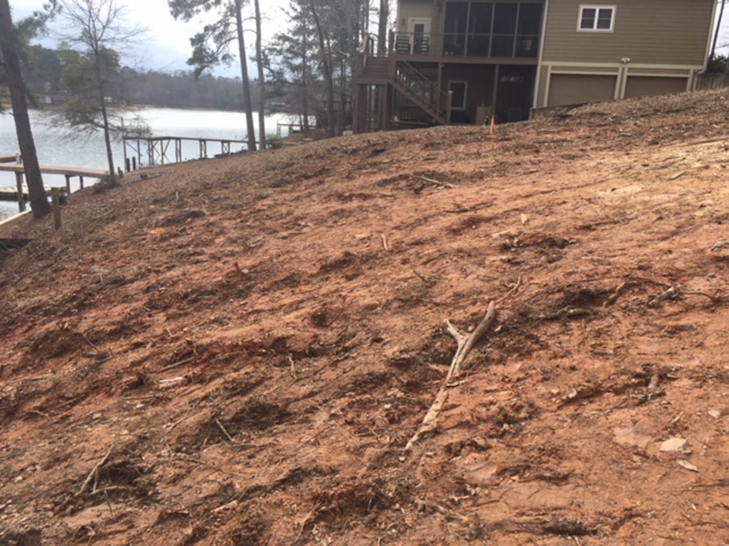 Two-story home with white-framed windows and garage door, situated on a dirt hill overlooking a lake, surrounded by trees and plants.