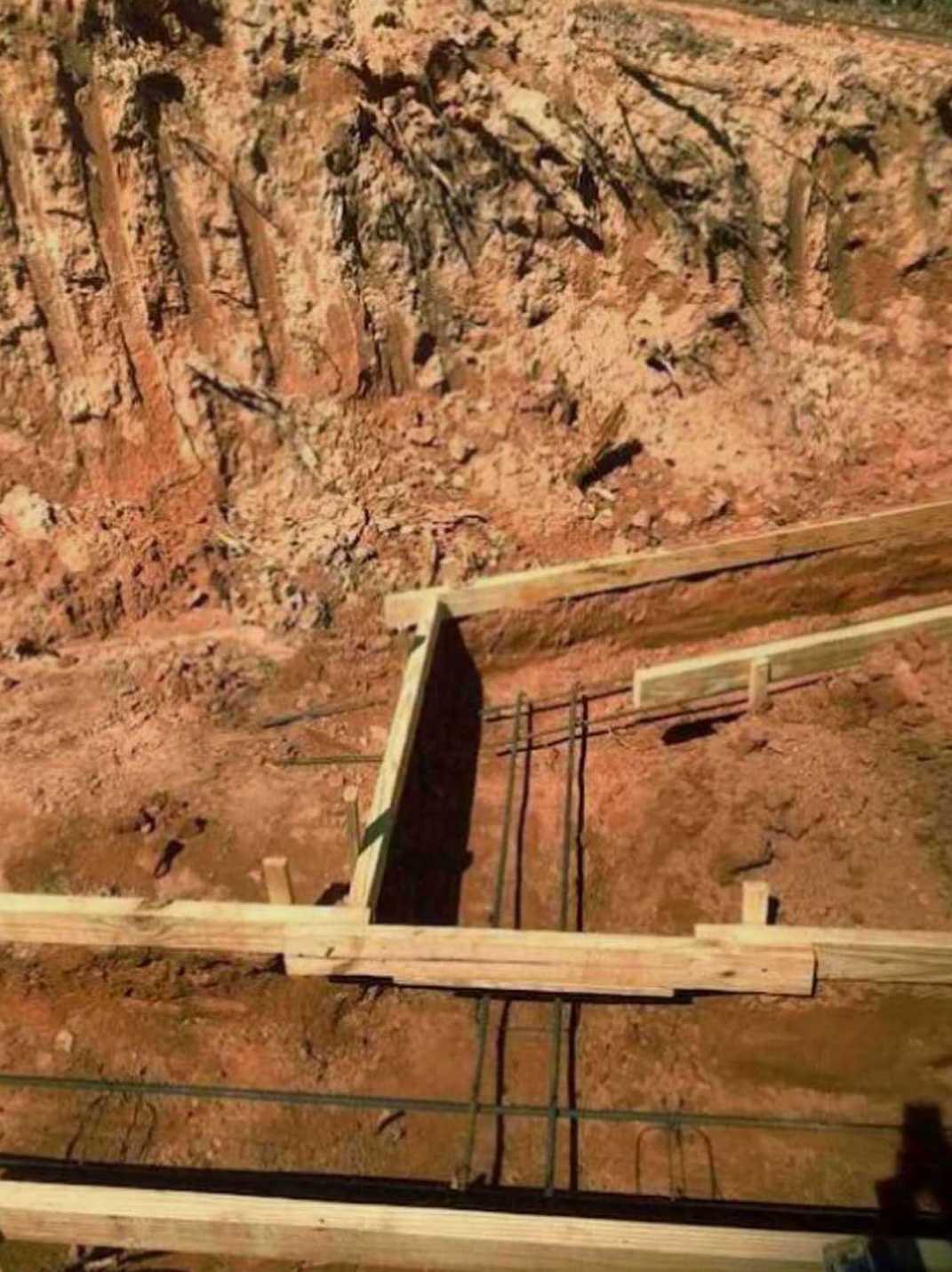 Wooden framing structure set on bare dirt ground outdoors, surrounded by rocky soil and construction materials