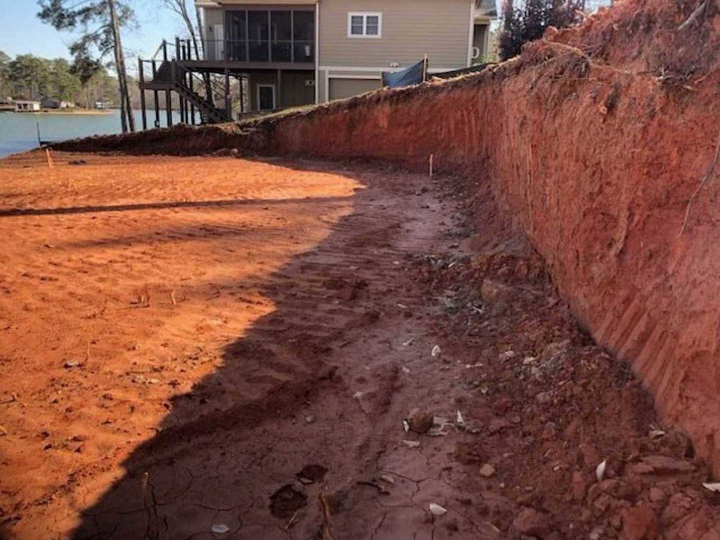 Dirt road bordered by trees leading to a modern house with white-framed windows, metal stairs, and fenced yard in the background