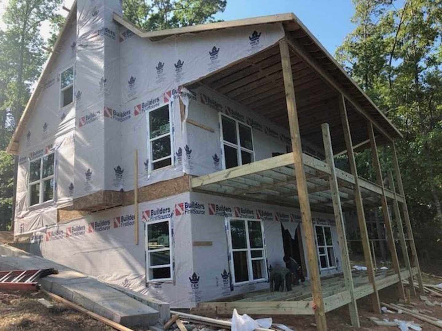 Two-story house under construction with exposed lumber framing, white window frames, scaffolding along exterior wall, and a dog standing on dirt in front yard