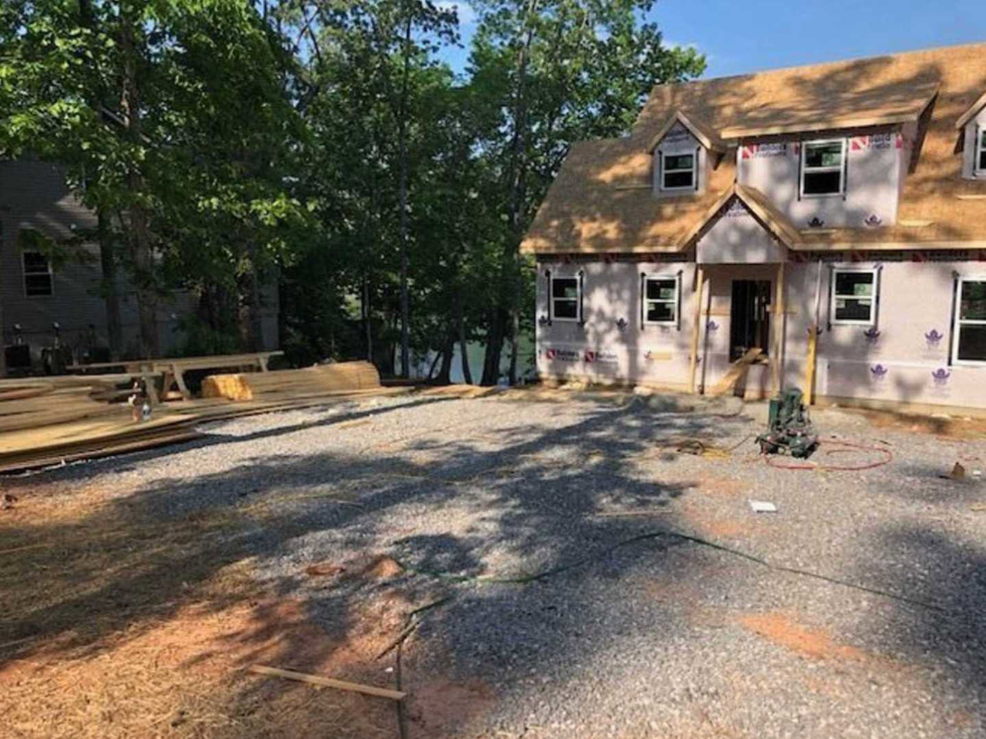 Framed house under construction with exposed plywood walls, white-trimmed windows, gravel driveway, and mature trees in the background