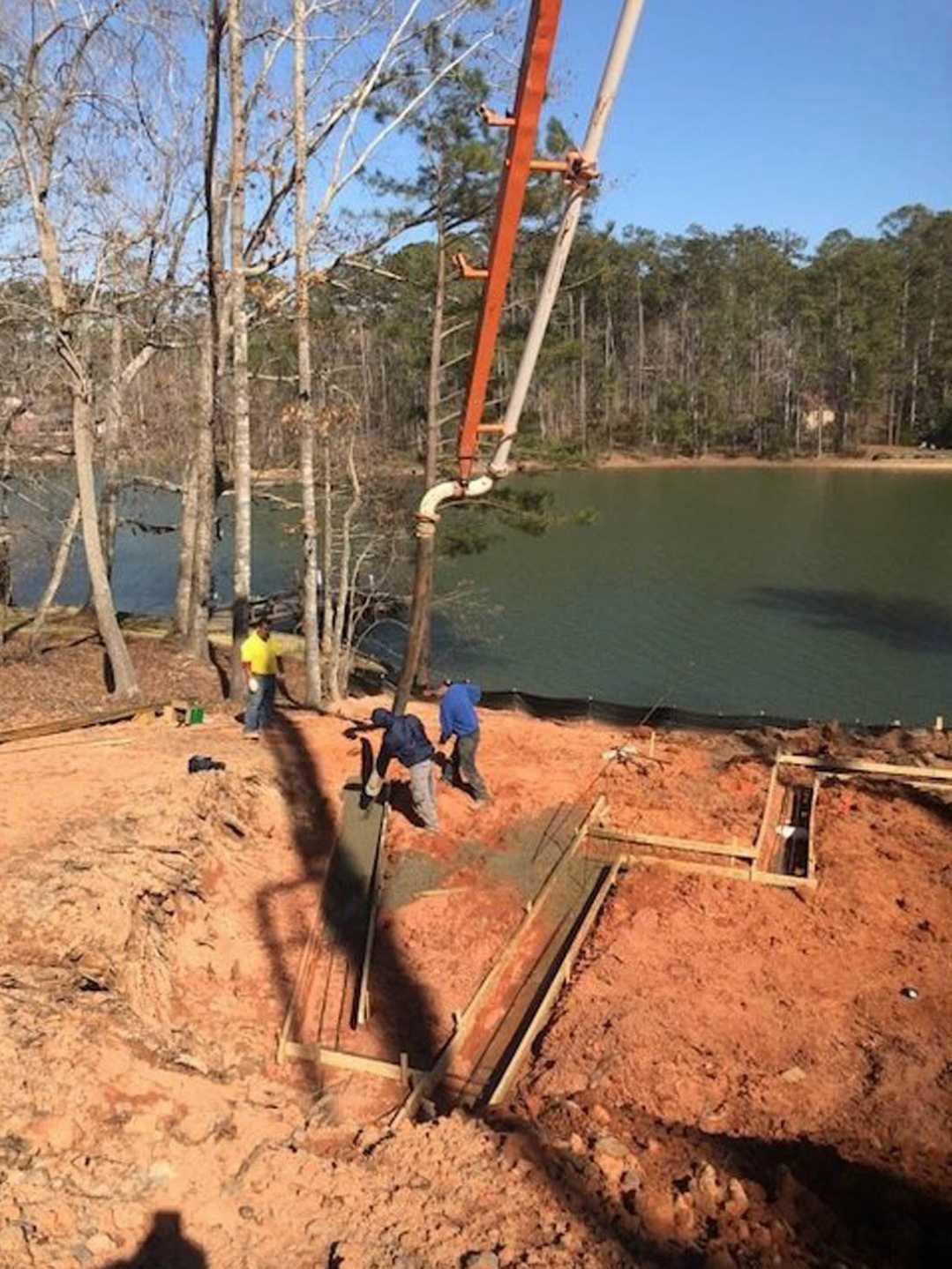 Workers assembling materials on a residential construction site with exposed framing, dirt ground, and scattered tools; trees and lake visible in background.