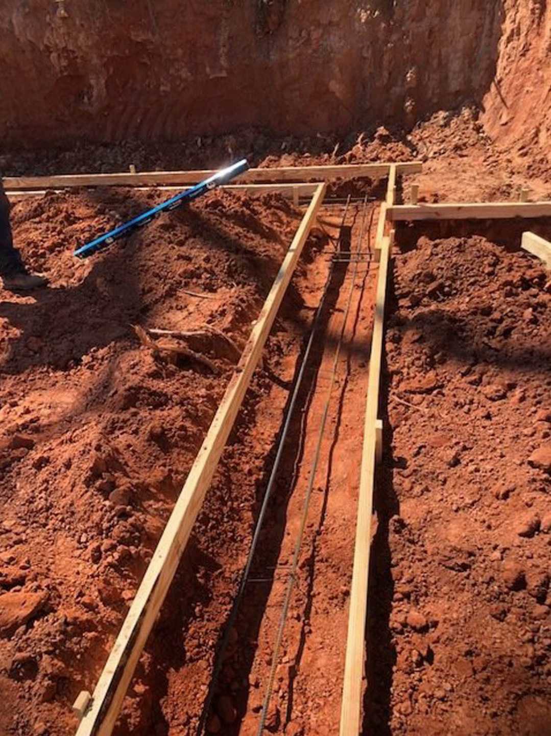 Man holding blue tool on dirt ground in front of unfinished wooden house frame