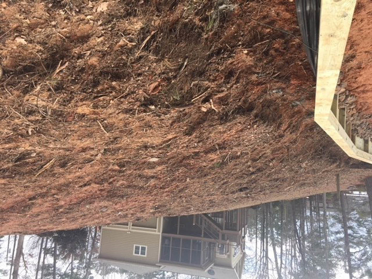 Two-story house with white-framed multi-pane windows and covered porch, perched atop a dirt hill with scattered trees; wooden bridge spans a cliff in the foreground.
