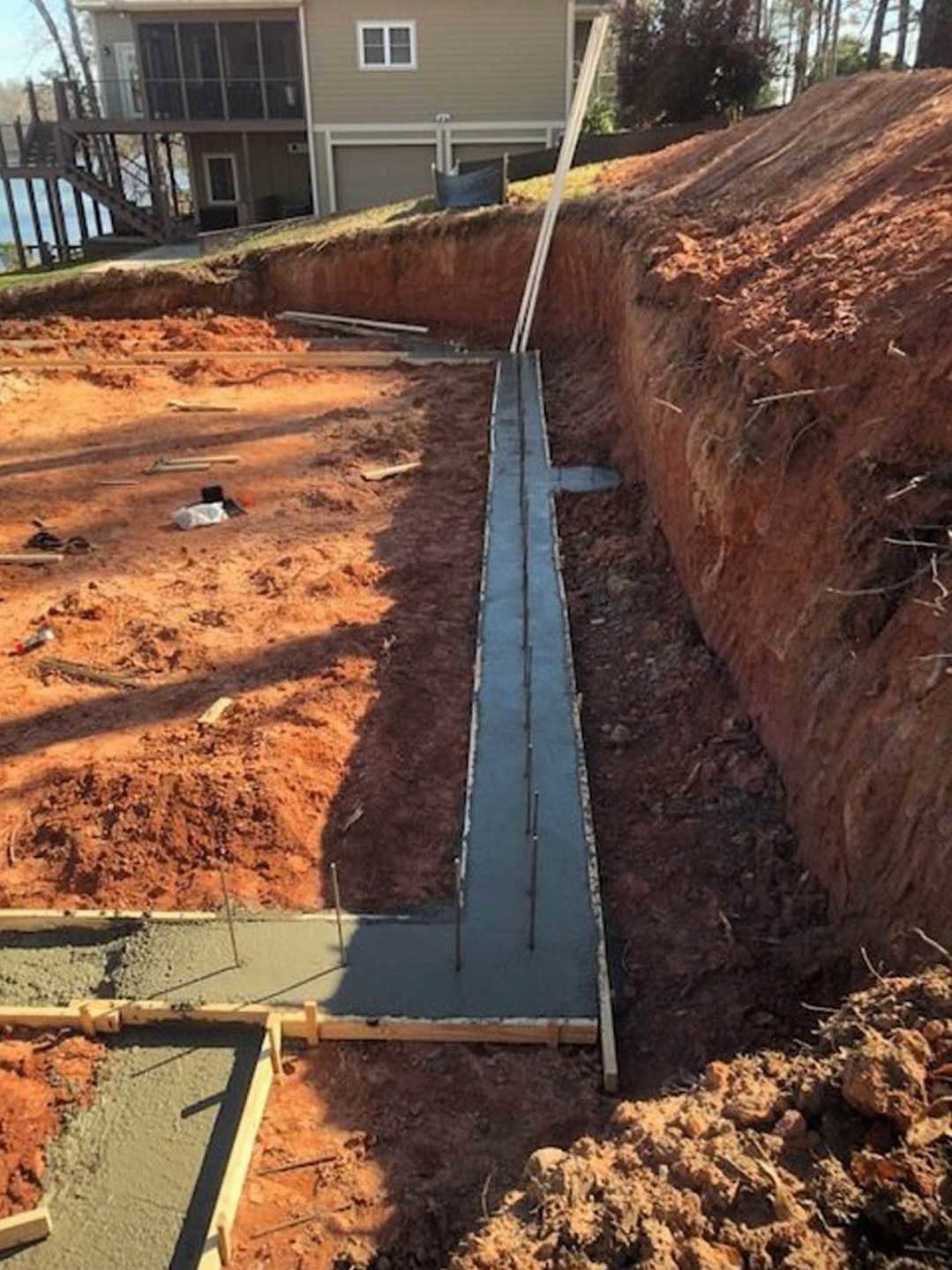 Two-story custom home under construction with exposed wood framing, white-framed windows, concrete foundation, pile of dirt in foreground, metal fence with blue band, and trees in
