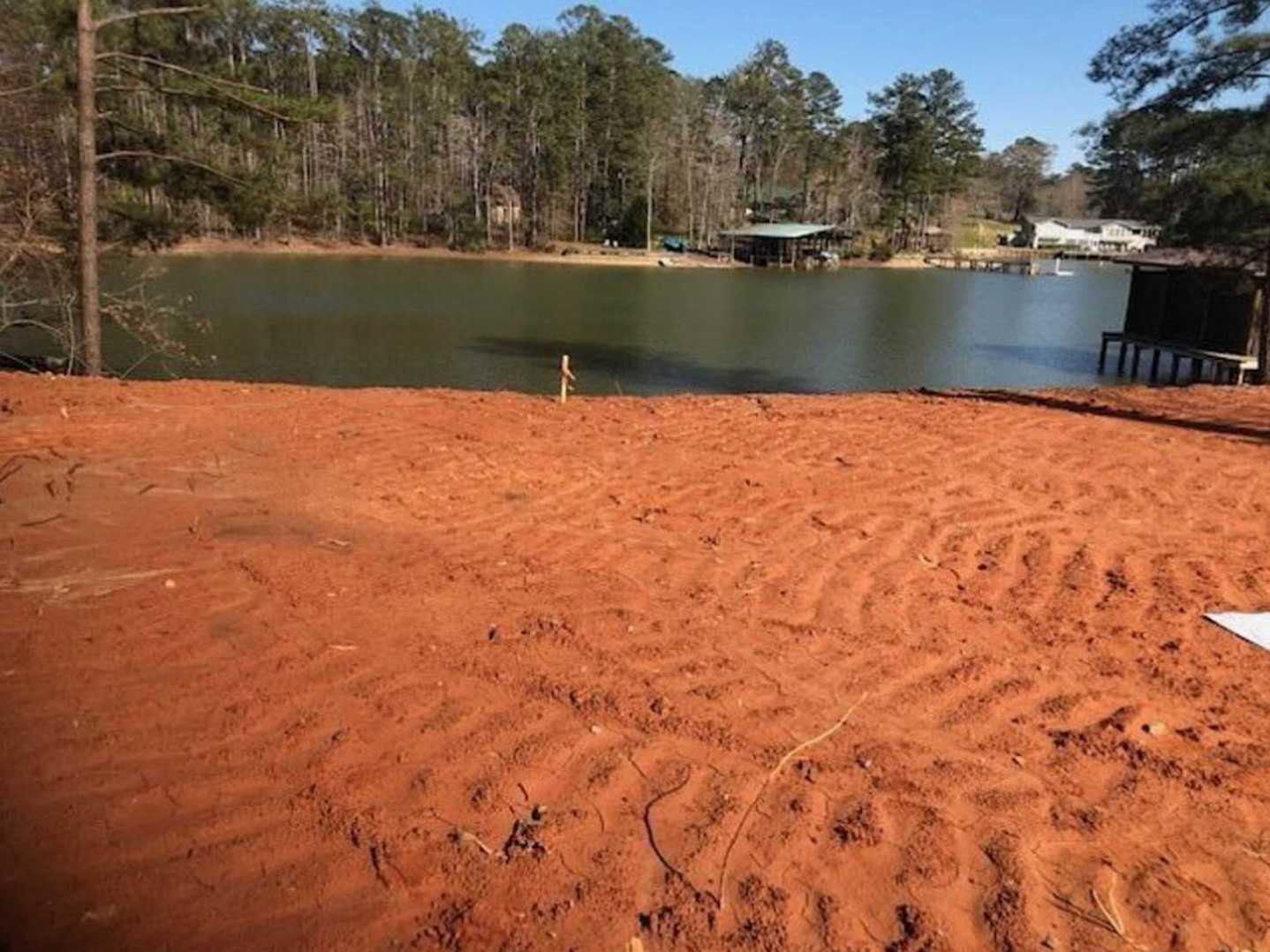Red sand beach bordered by trees, calm lake water, and clear sky in the background.