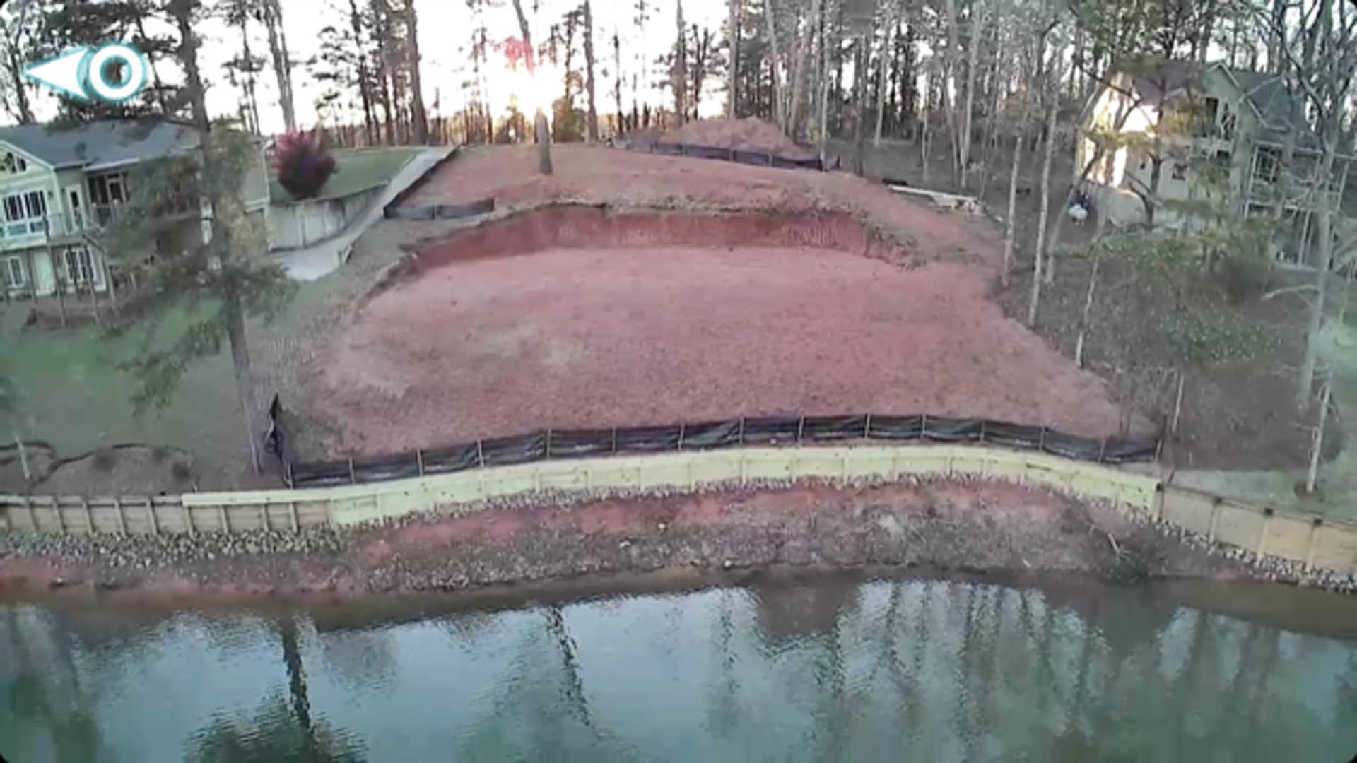 Pond bordered by a dirt hill and mature trees, reflecting greenery and sky on calm water