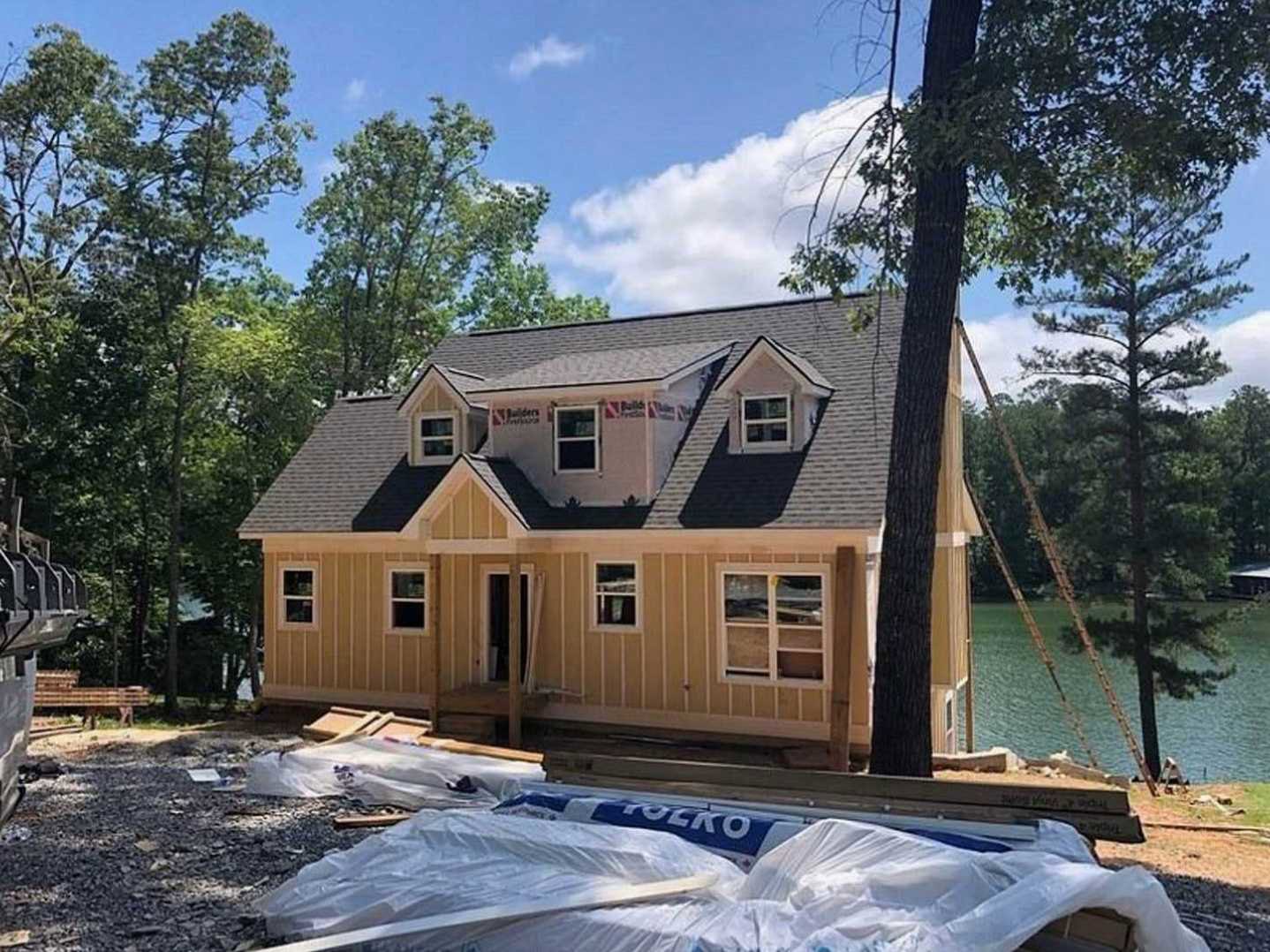 Framed house under construction with exposed wood beams, surrounded by tall trees, lake visible in background, cloudy sky overhead