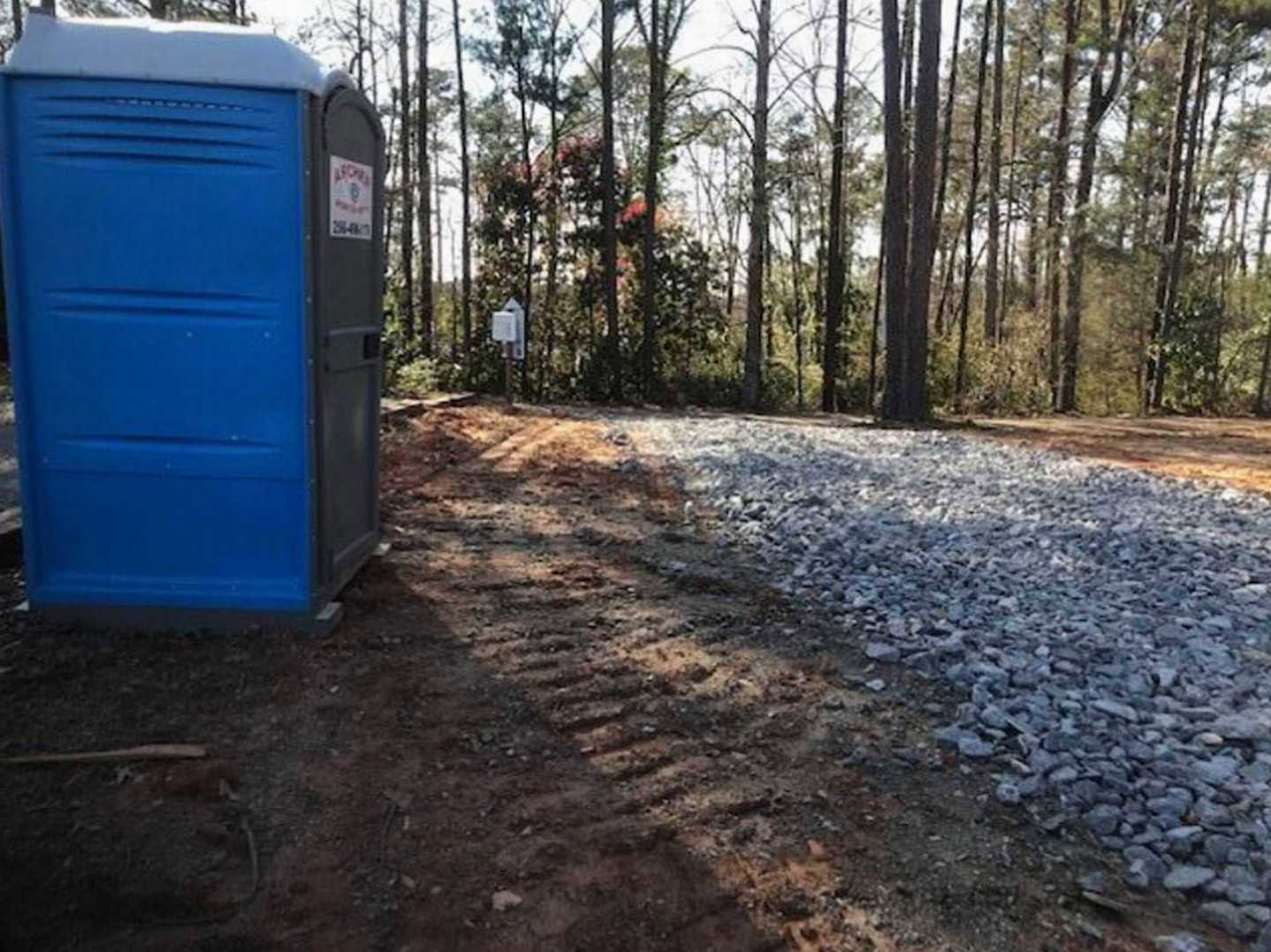 Blue portable toilet set beside large rocks and tall trees on gravel ground, with forested background and clear sky