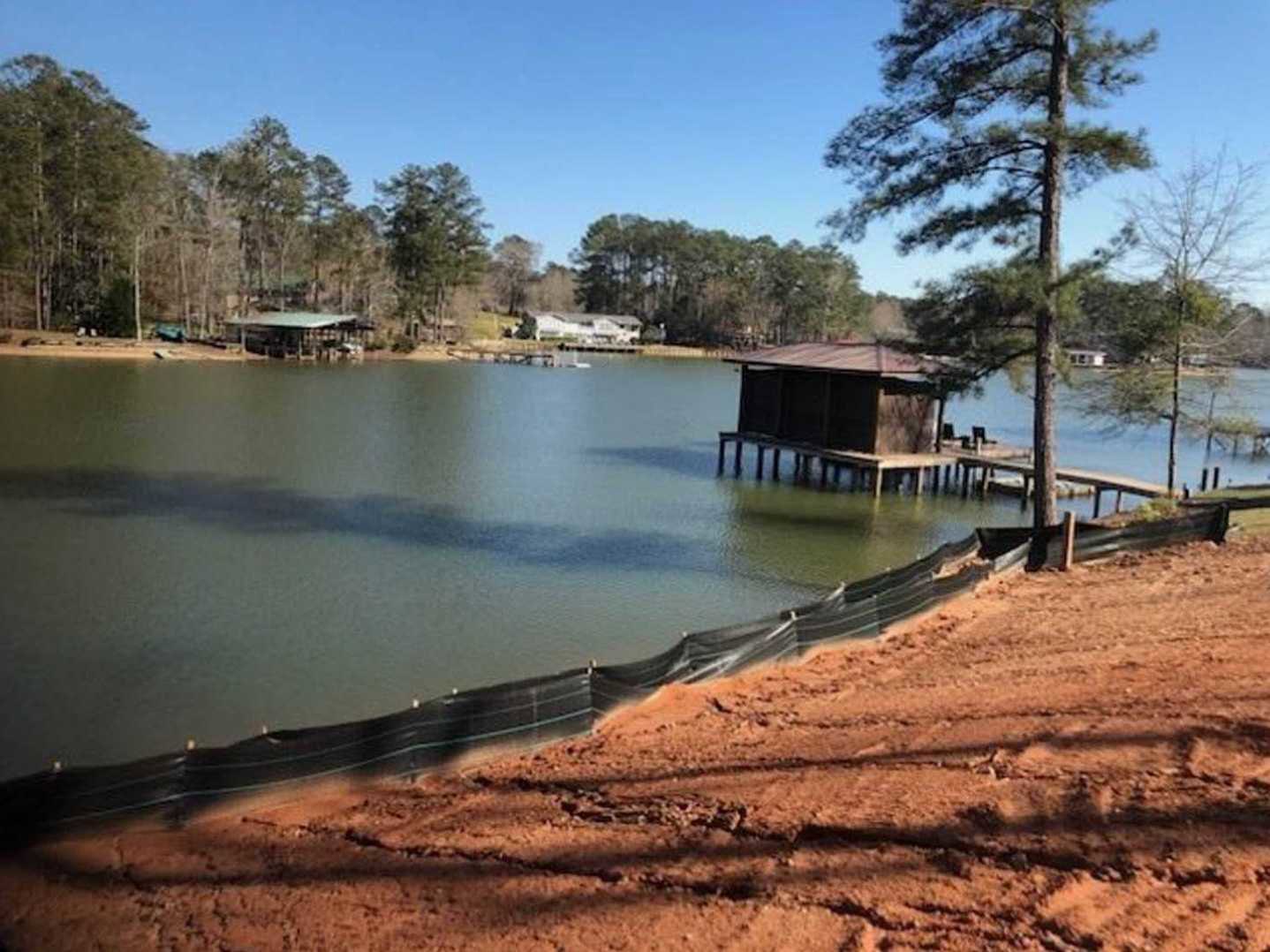Small wooden cabin elevated on stilts over a calm lake, sandy shoreline bordered by a black metal fence, leafy tree beside the structure, blue sky and dense trees in the background