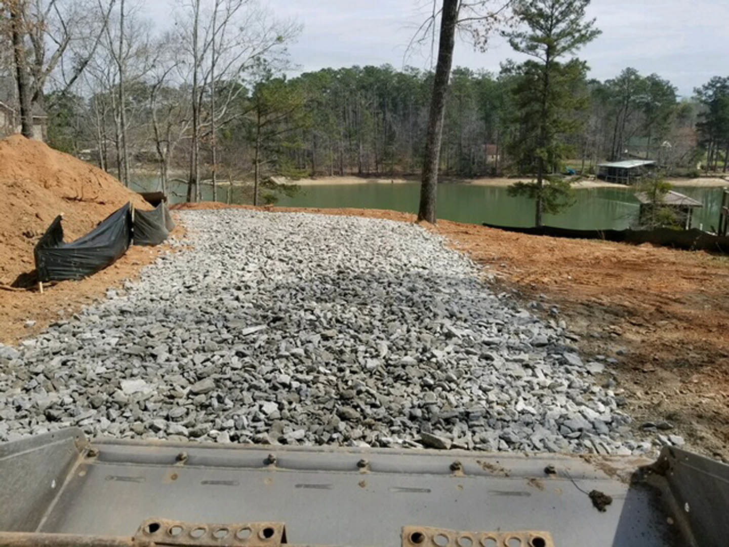 Rock pile on sloped dirt hill with black tarp, surrounded by green trees and lake in background, under open sky.