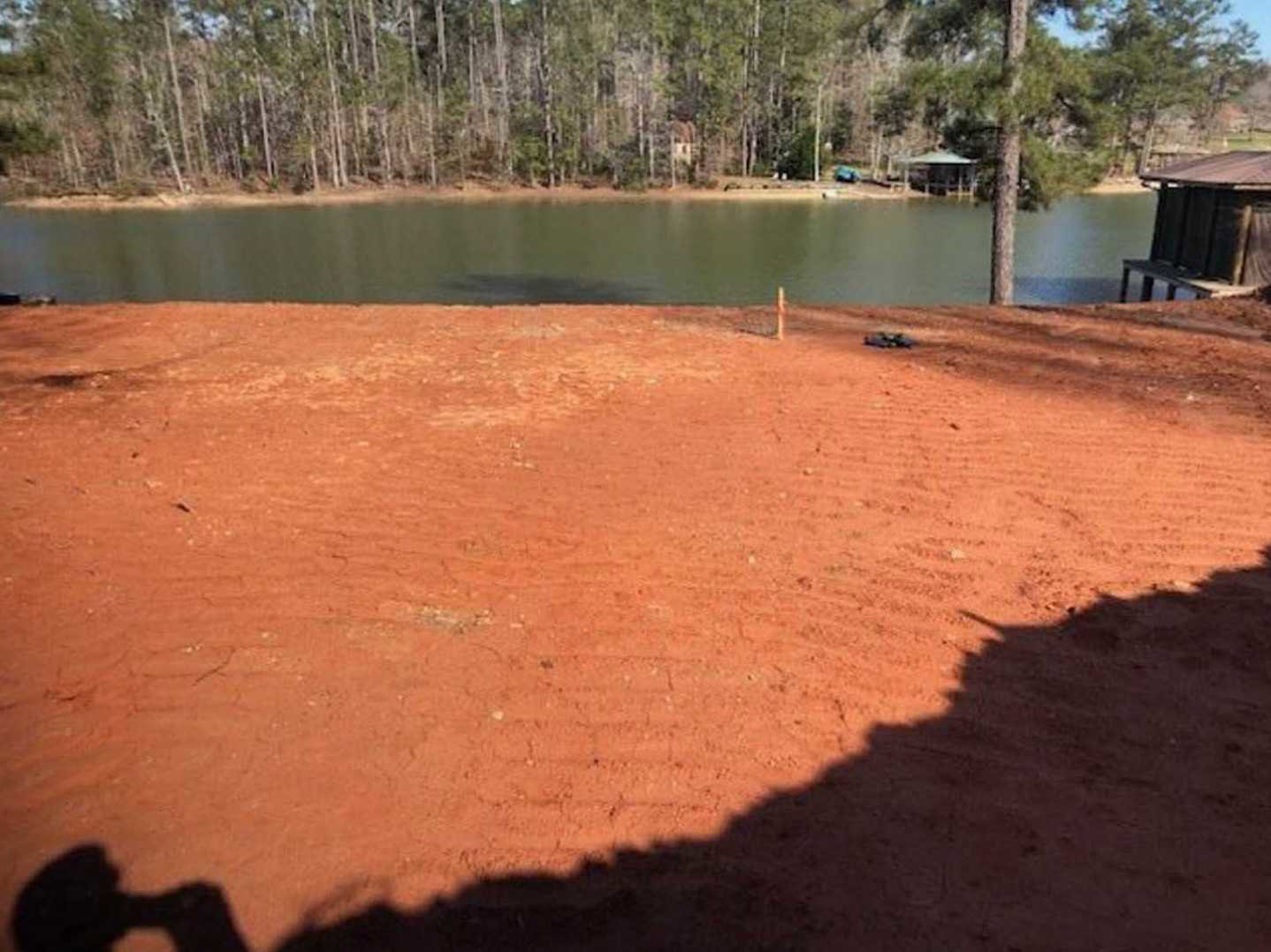 Red dirt field bordered by trees, wooden fence, and a small structure beside a lake, with a person standing near the water