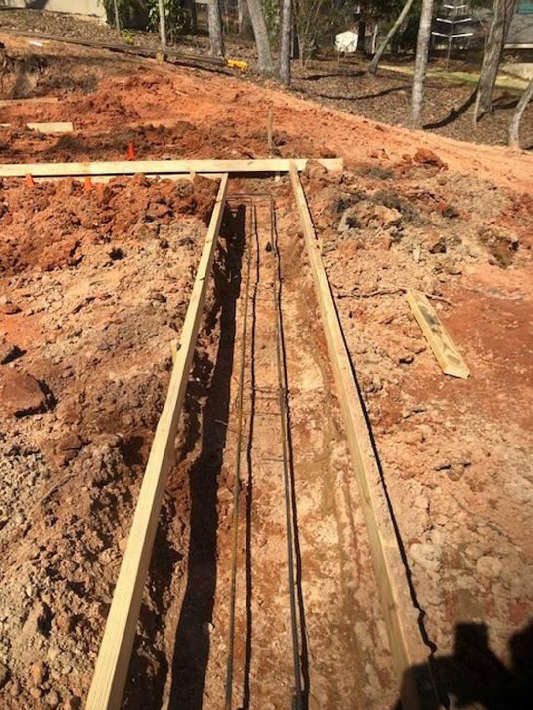Wooden support beam resting on soil at residential construction site, surrounded by dirt and scattered rocks, with blurred trees and window in background