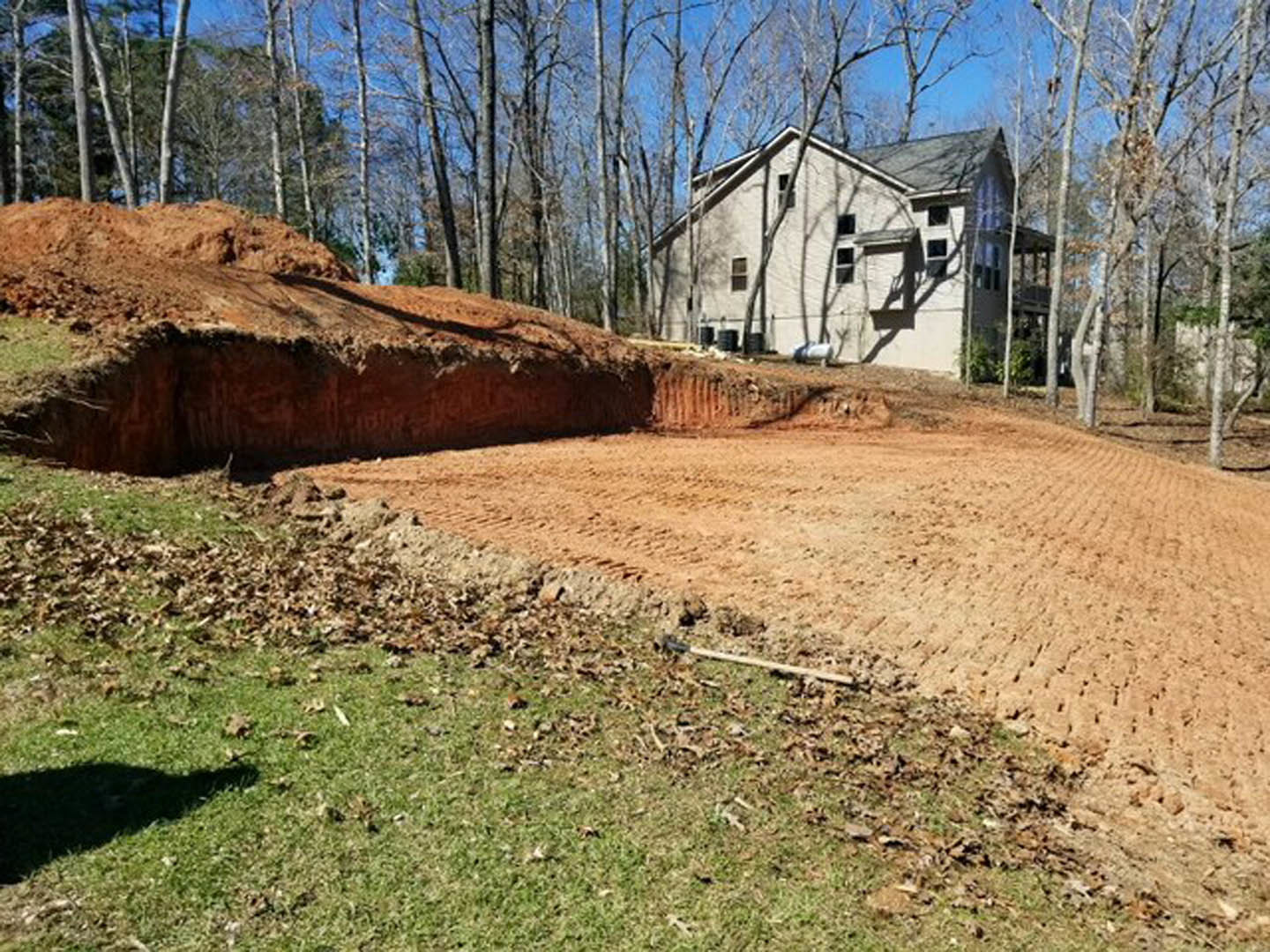 Modern house with light siding and dark roof partially visible behind a dirt hill, surrounded by mature trees and grassy lawn, shovel resting on soil in foreground.