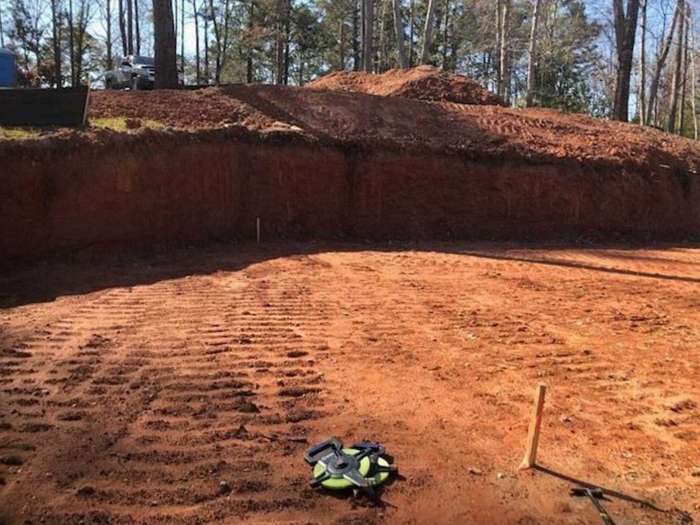Dirt pit with a green and black disc lying on the ground, hammer and pole nearby, dirt hill and trees in the background, parked car partially visible next to a tree.