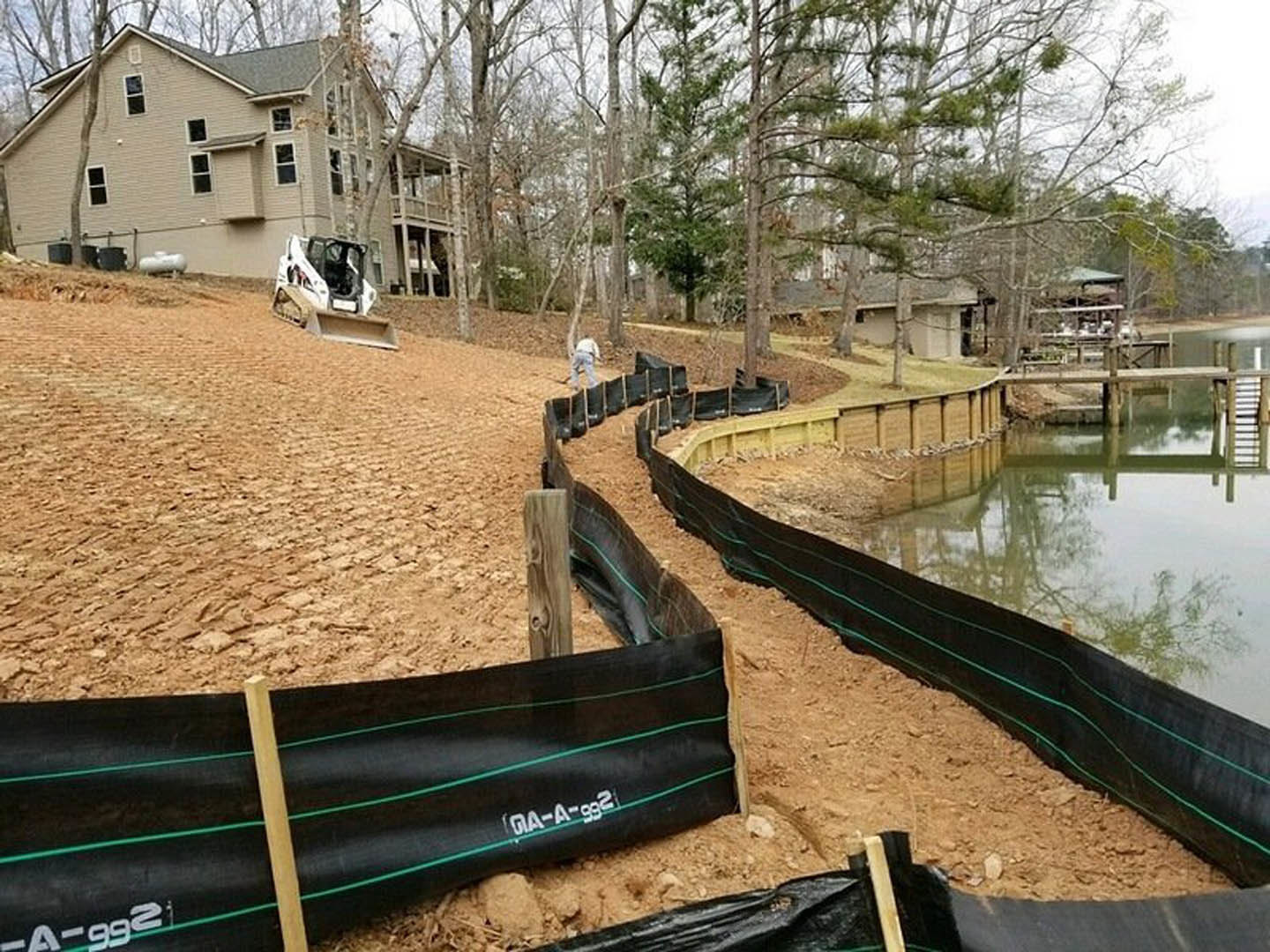 Bulldozer and tractor parked near a partially constructed house beside a pond, black plastic tubing and wooden bridge visible, surrounded by trees and grassy ground.