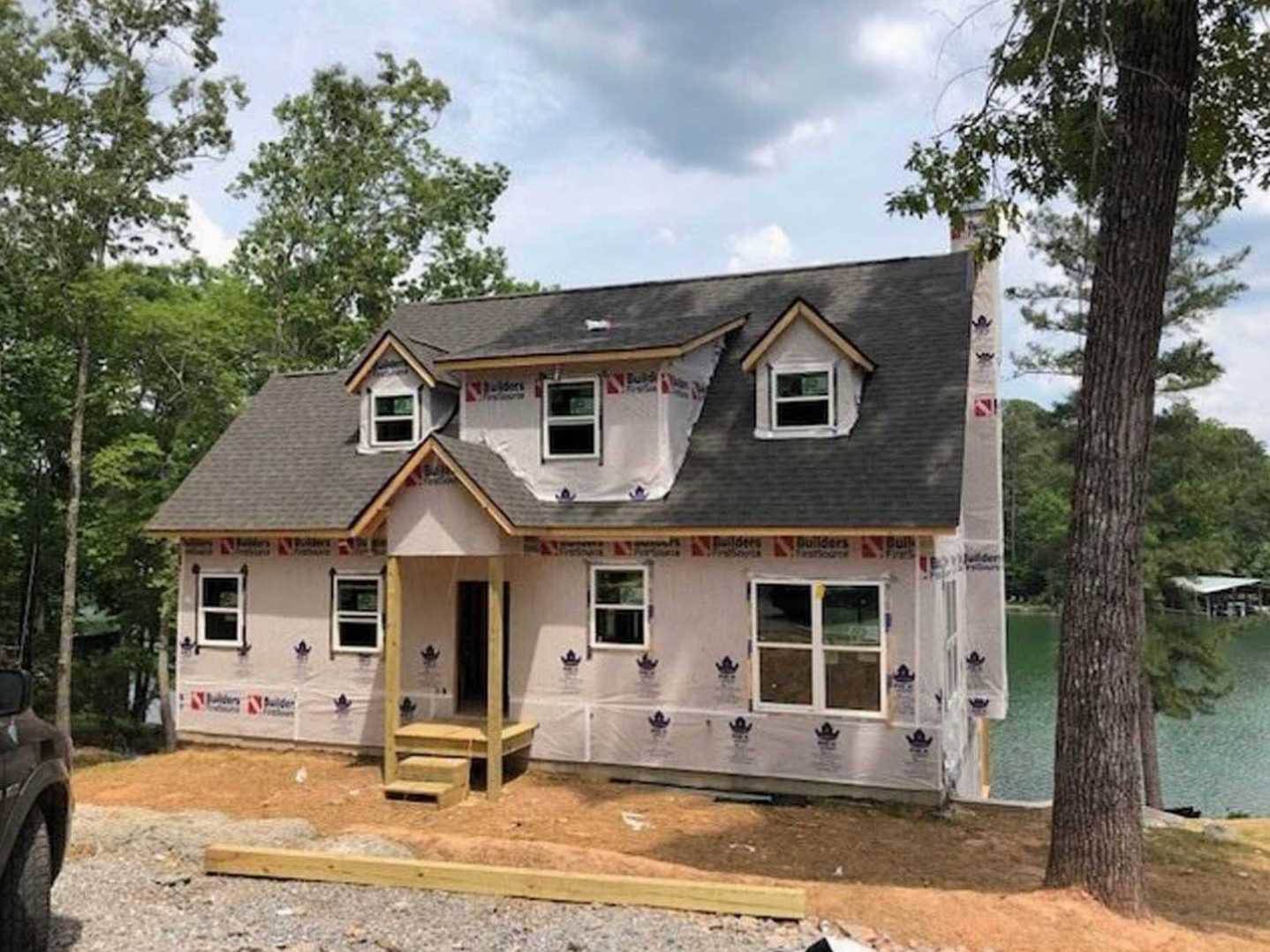 Partially built house with plastic sheeting, white-framed window, tree trunks nearby, and lake visible in the background