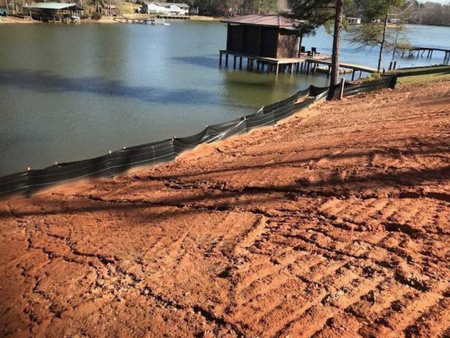 Dirt hill with black plastic barrier fence, small stilted building, dock with chairs, body of water, tree trunk in foreground