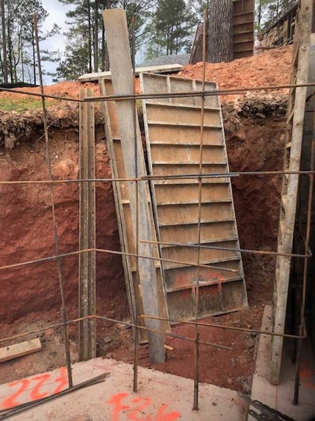 Stack of wood planks and metal bars in a dirt excavation at a residential construction site, surrounded by soil and unfinished building materials