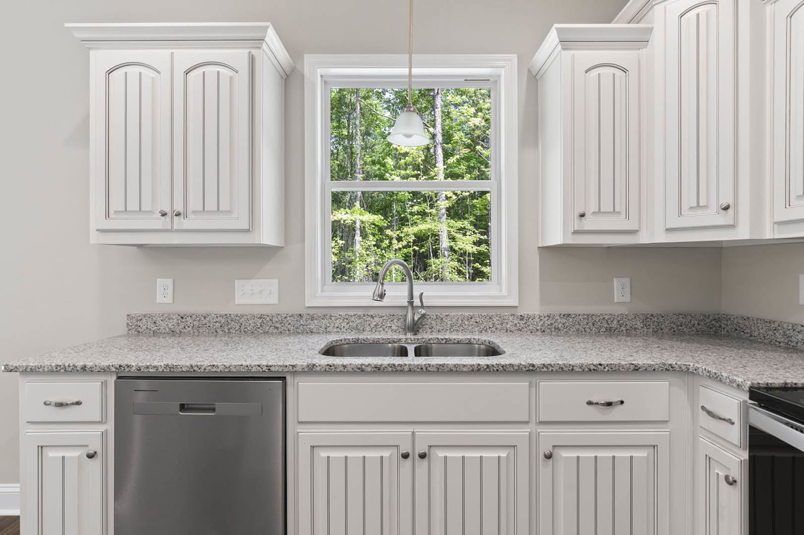 White kitchen cabinets with silver knobs, stainless steel dishwasher, stone countertop, chrome faucet, and natural light from a window illuminating the sink area.