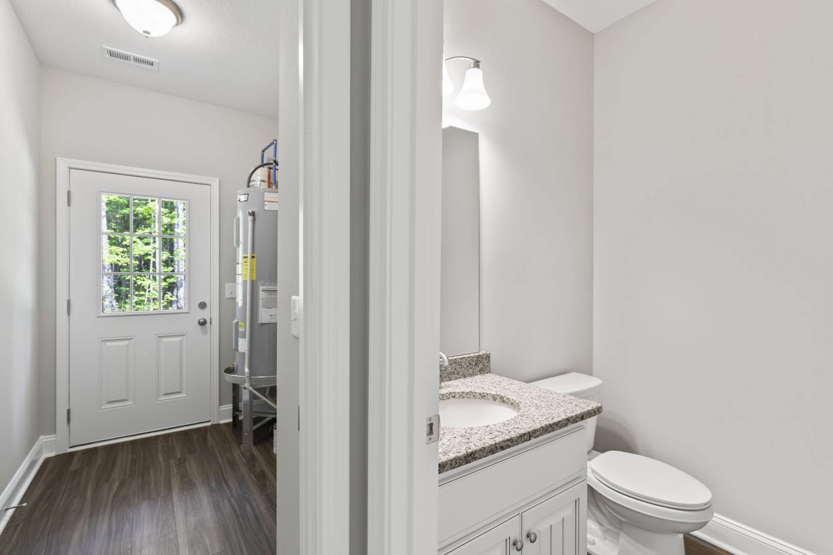 Bathroom with white toilet and sink, wood flooring, white door with glass window, tiled walls, and shower tub visible in background.