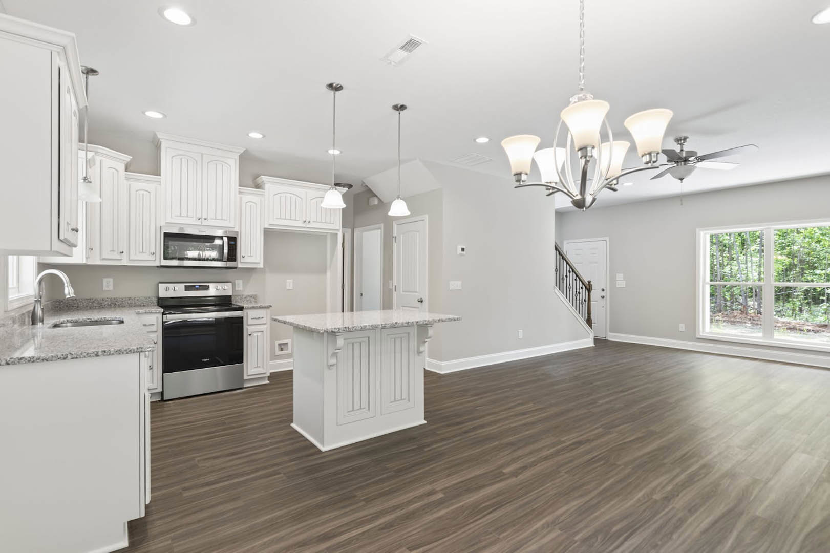 Open kitchen and dining area featuring hardwood flooring, white cabinetry, marble-topped island, stainless steel stove, and large window overlooking trees.