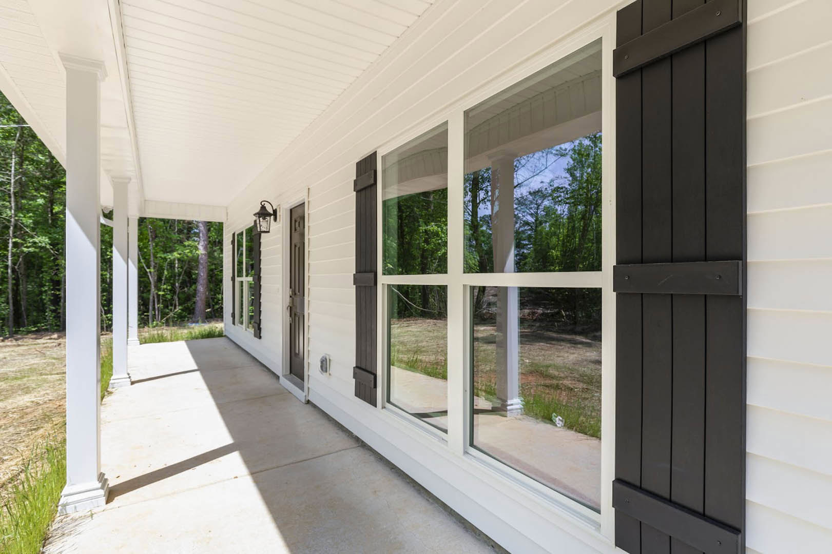 White siding home with black shutters, black front door, covered porch, windows, green lawn, and mature trees in the background