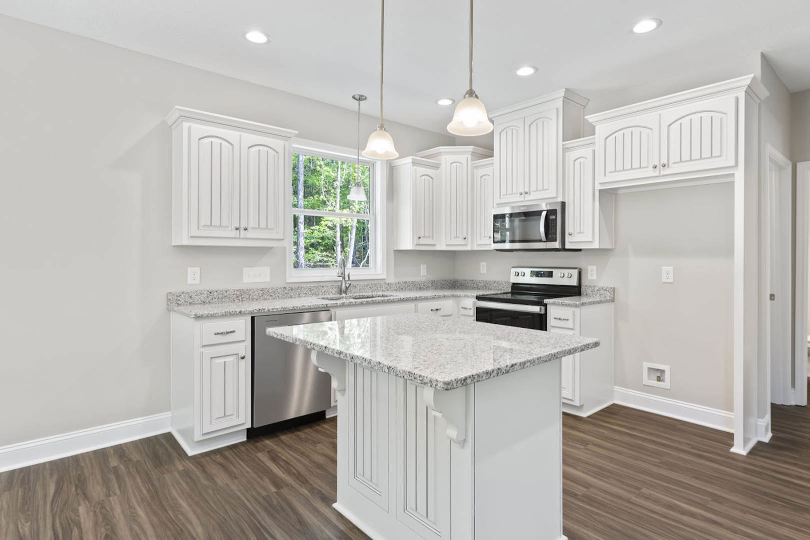 White kitchen with granite countertops, white cabinets, marble-topped island, stainless steel stove, built-in microwave, and window with white trim letting in natural light.