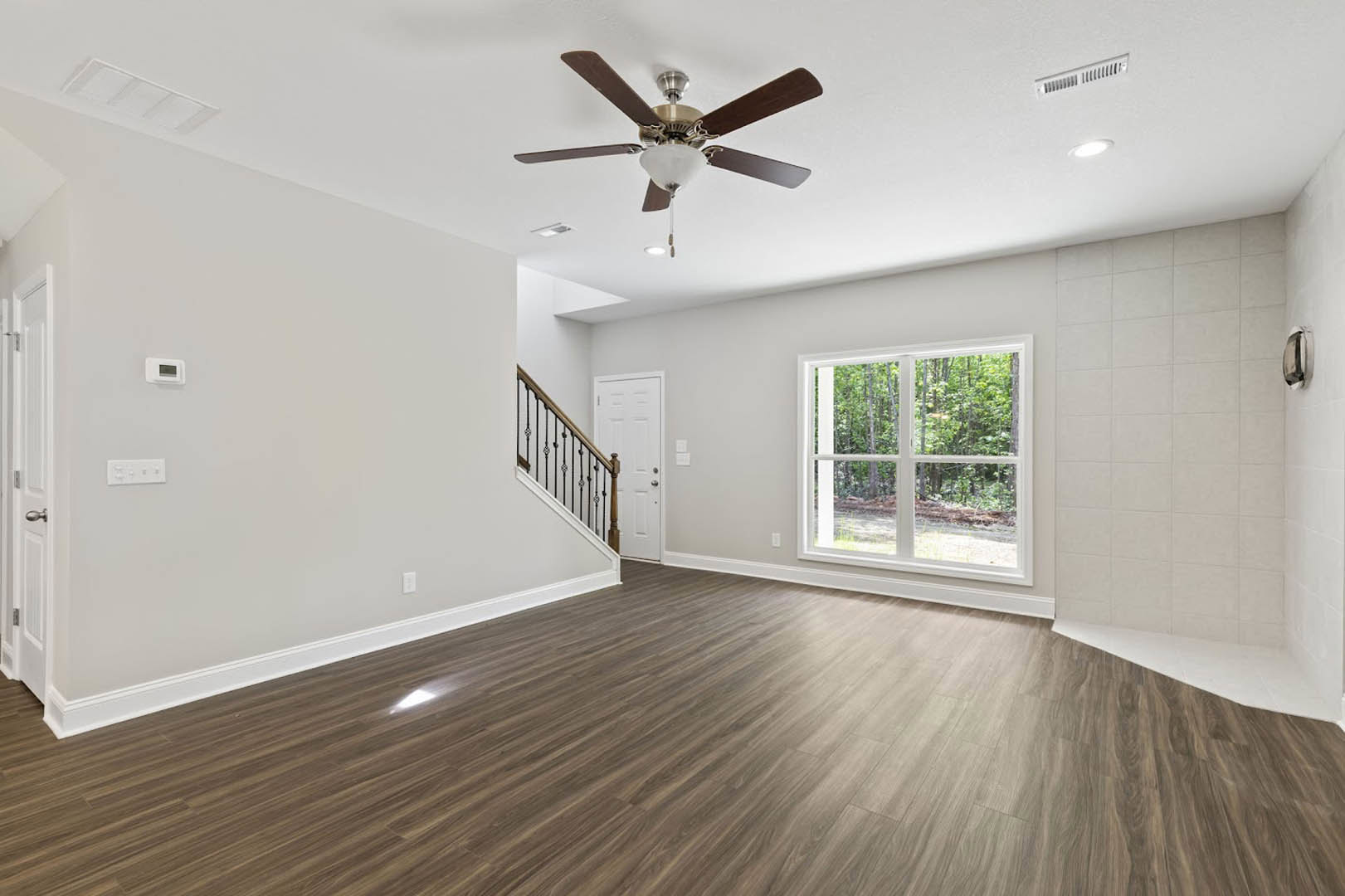 Hardwood floor room with white walls, ceiling fan and light fixture, window overlooking trees, close-up staircase, two white doors