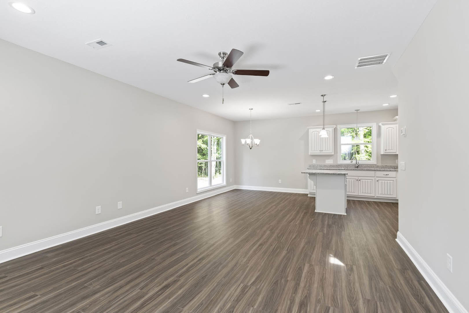 Open room with wood flooring, white walls, ceiling fan with light fixture, large window showing trees outside, wall vent, and window above a sink.