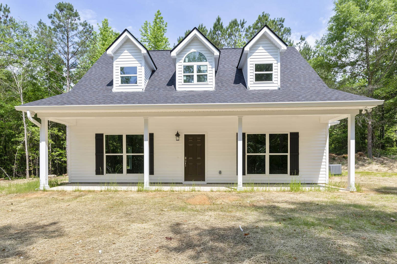 White siding house with black shingle roof, black front door, dormer windows, grassy lawn, and covered porch