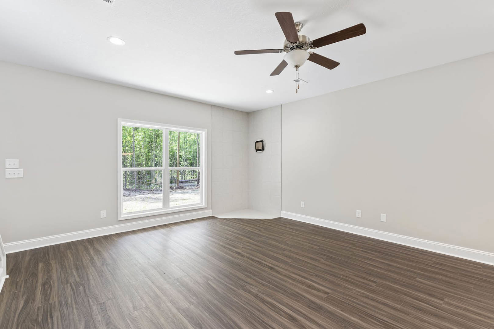 Ceiling fan with light fixture mounted on white ceiling, wood flooring, white walls, window overlooking trees, white light switch on wall