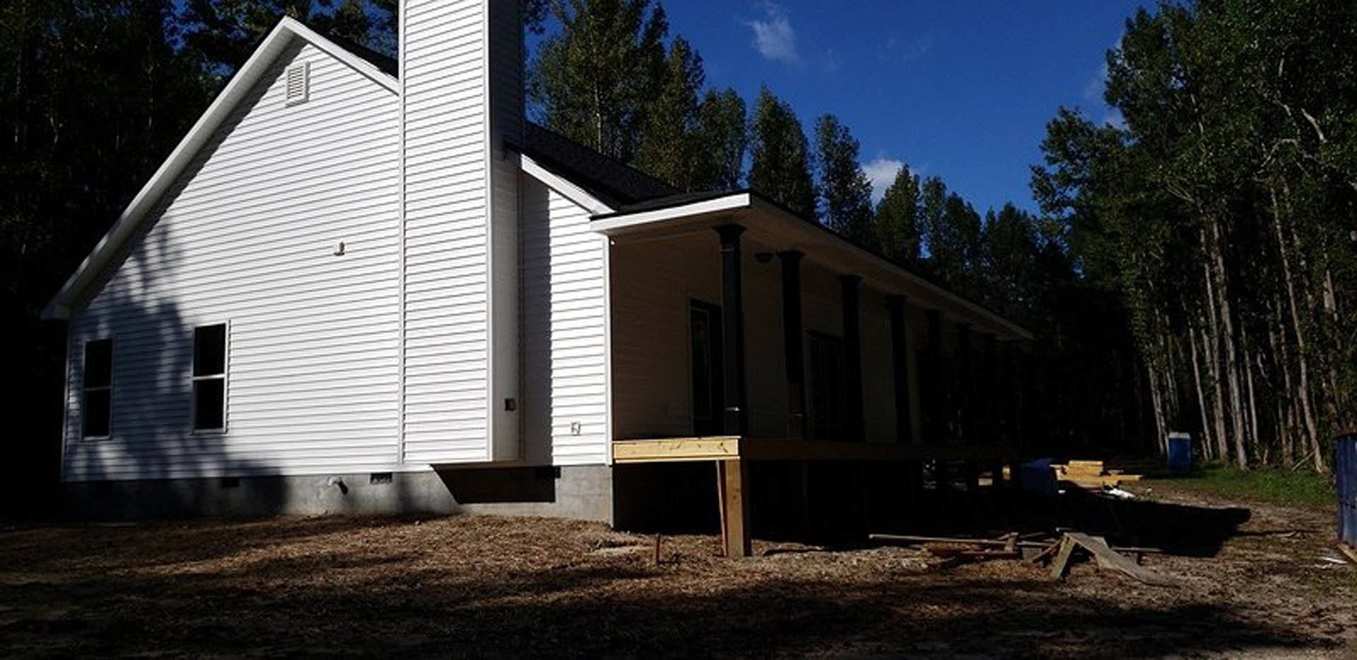 Wood-framed house under construction with exposed beams, unfinished siding, and large windows, surrounded by mature trees and clear sky in the background