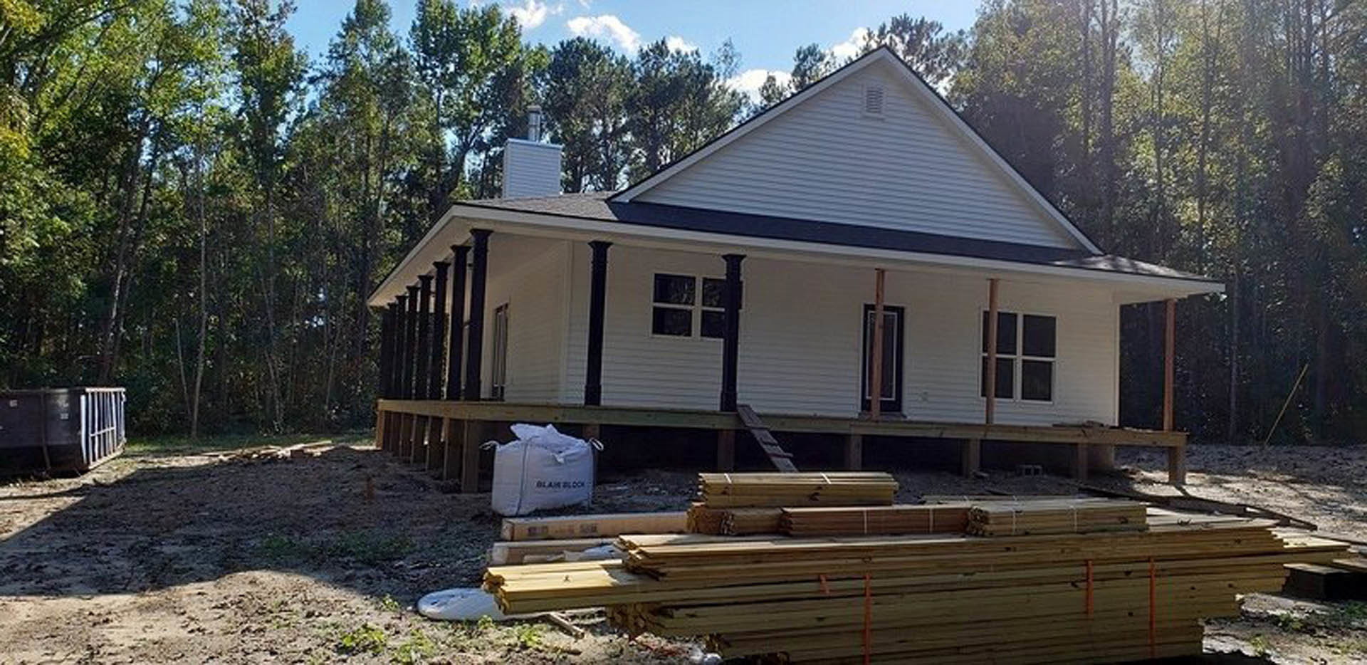 Wood-framed house under construction with exposed lumber, partially built porch, and stacks of planks in the yard; siding and windows not yet installed, surrounded by trees and