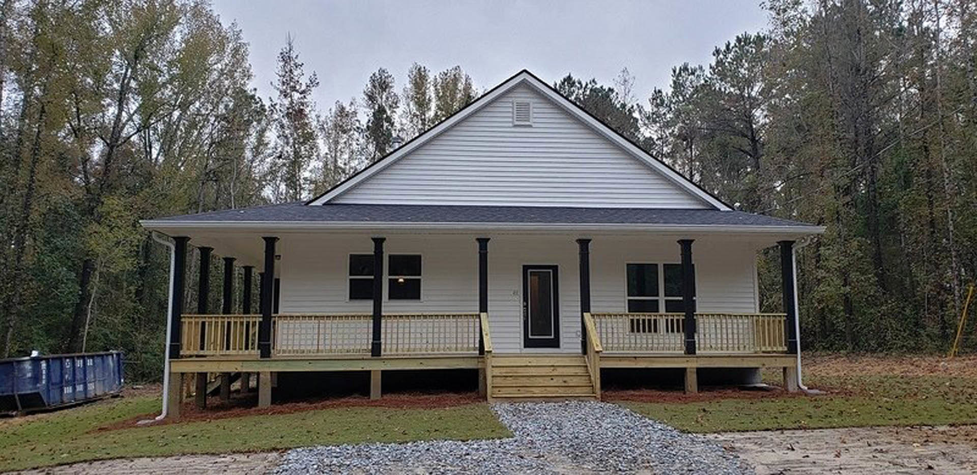 Two-story house with white siding, covered front porch with wooden columns, concrete driveway, landscaped yard with shrubs and trees, large windows, and gabled roof