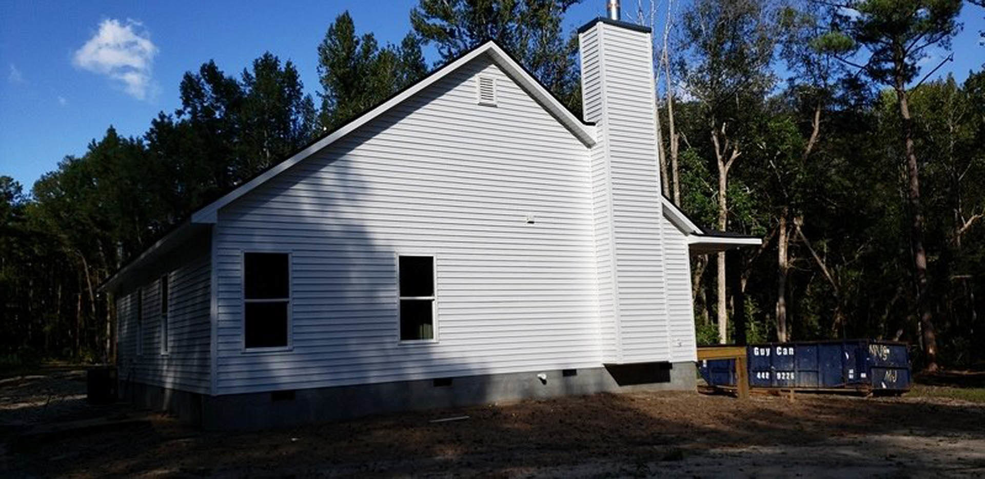 White siding house with brick chimney, black framed windows, surrounded by trees under cloudy sky