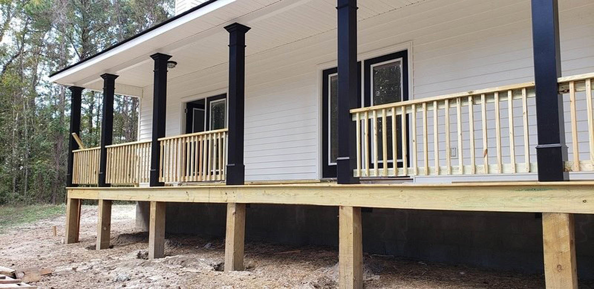 Two-story house with wood siding, covered porch supported by pillars, wooden railings, upper balcony, and mature trees in the yard