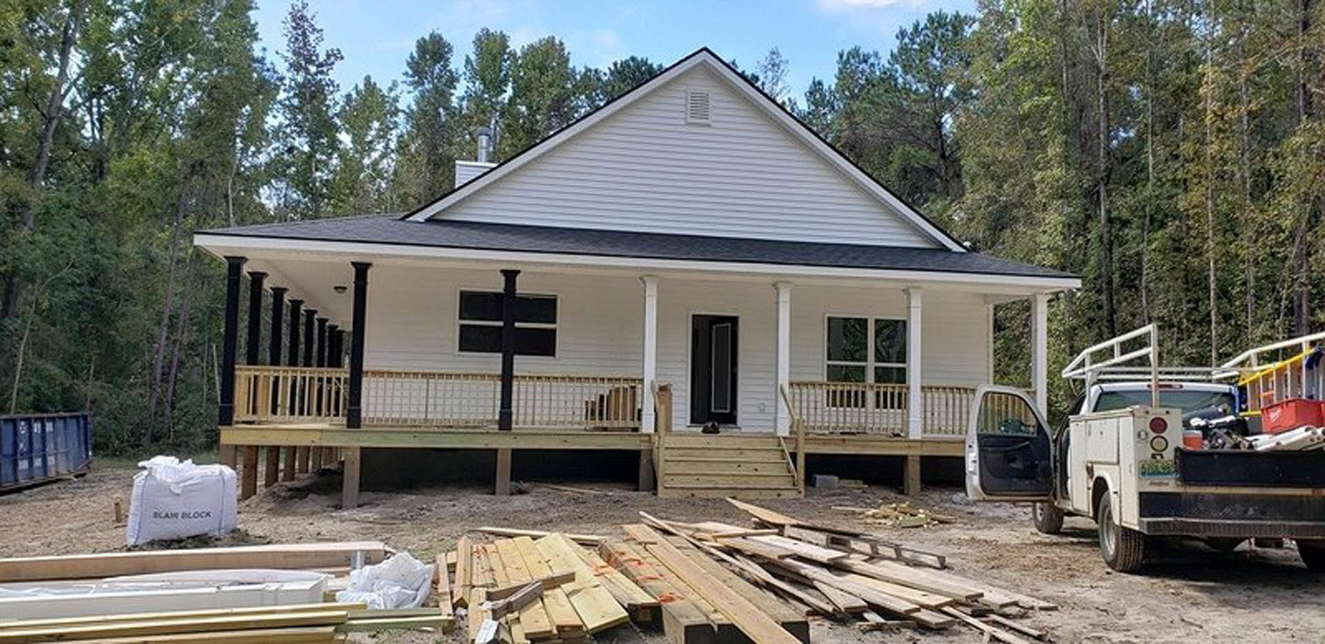 Two-story custom home with wood siding and trim, covered front porch with wooden railing, white work truck parked in driveway, construction materials and lumber stacked near