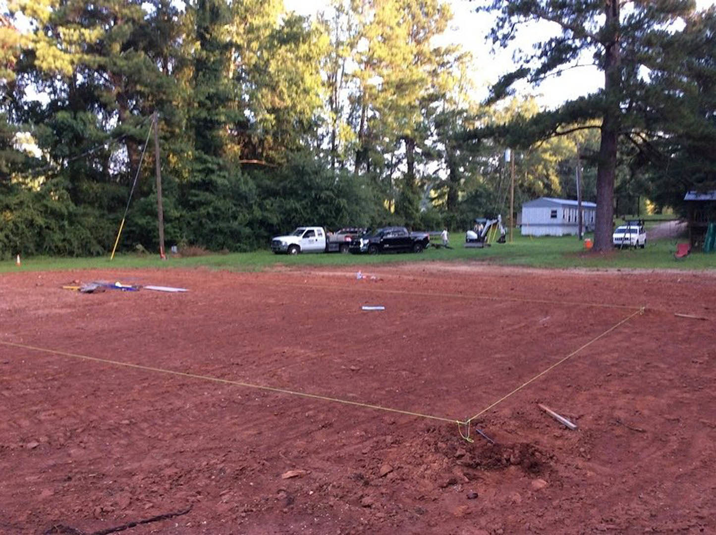 Red dirt field bordered by trees, white truck parked near the edge, sparse grass and plants scattered across the ground