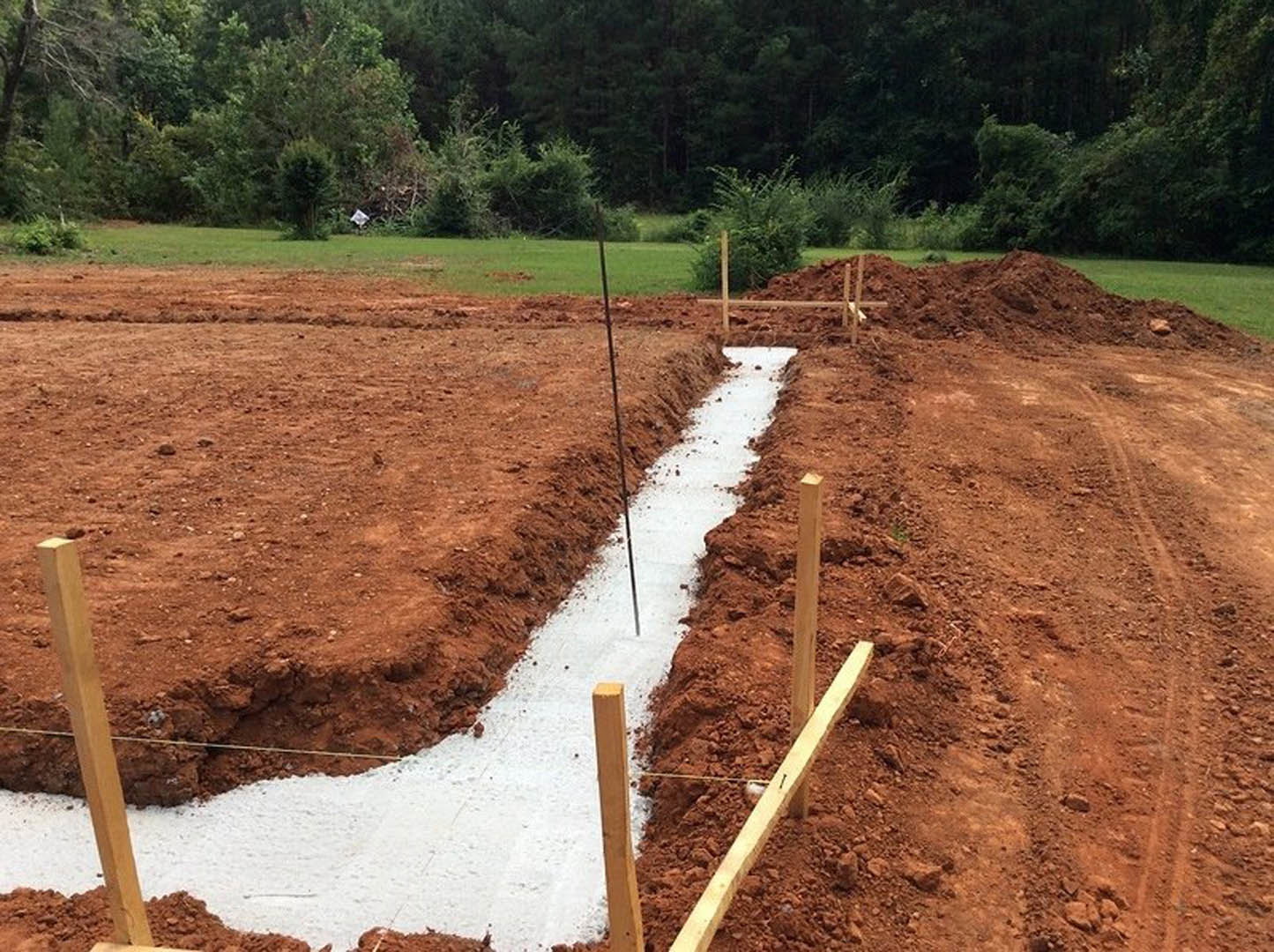 Concrete foundation slab surrounded by dirt and wooden posts, bordered by grass, trees, and bushes on a residential construction site