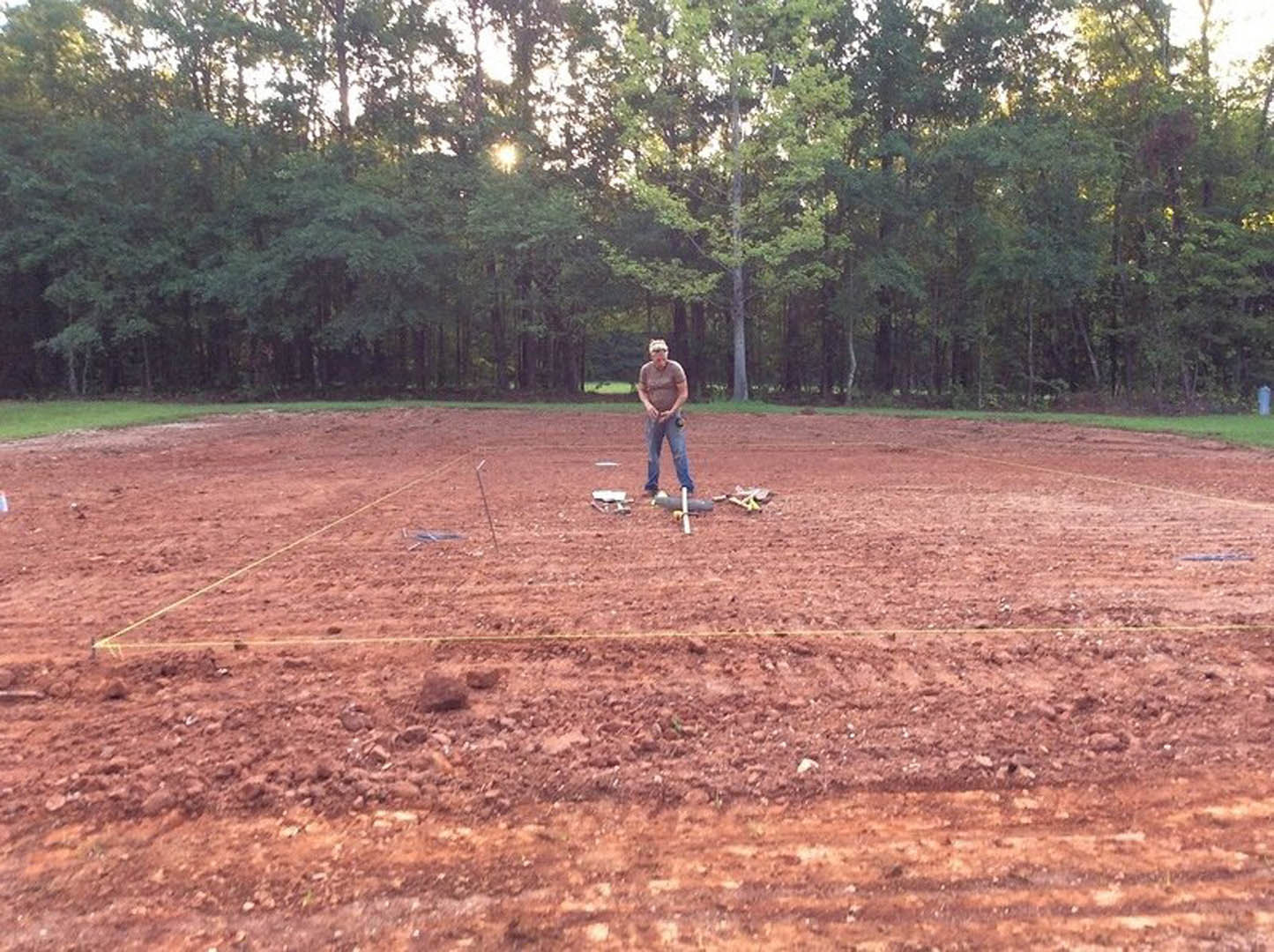 Man in blue jeans standing in a dirt field with scattered grass and trees in the background