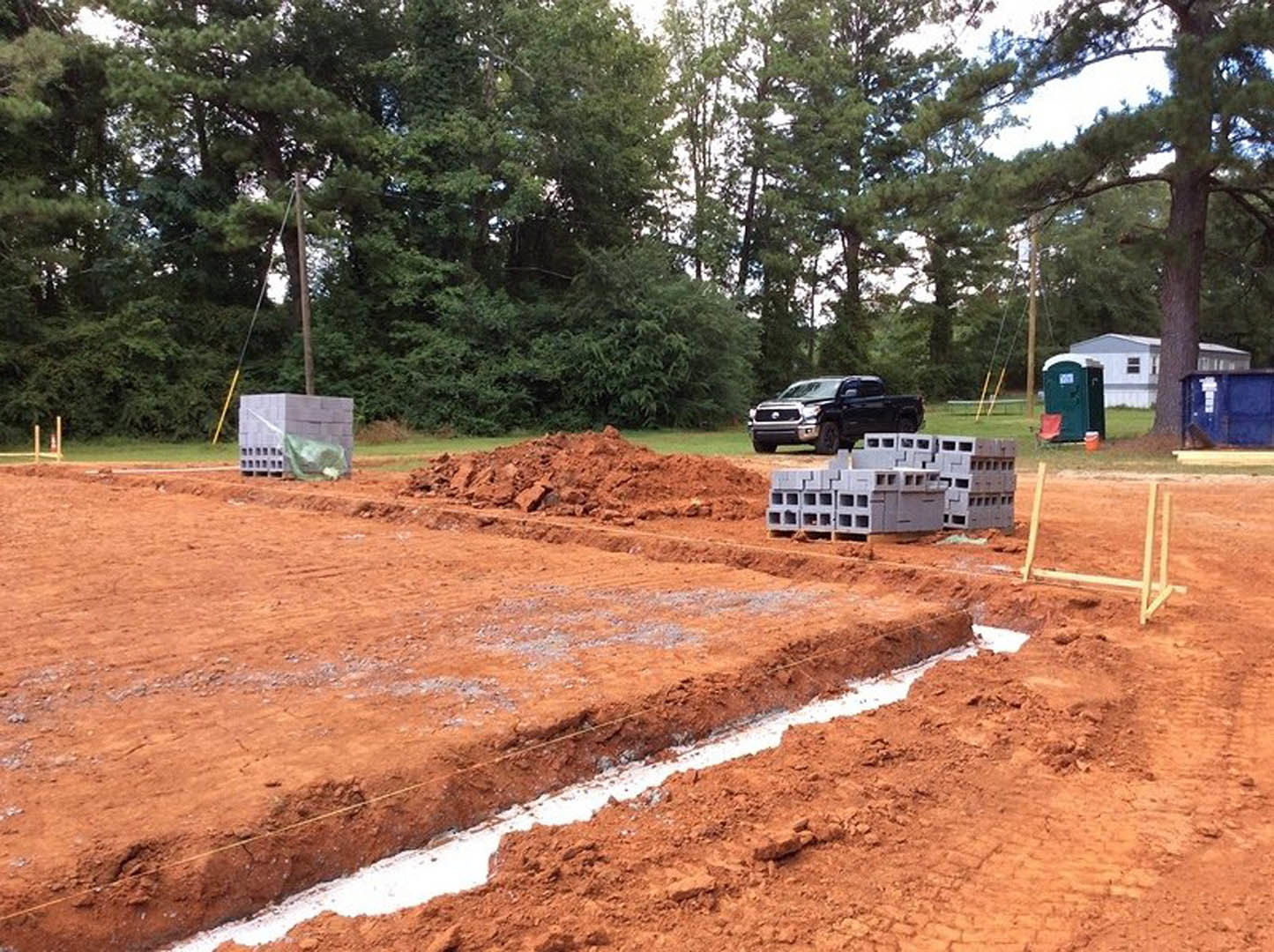 Partially built custom home with exposed concrete block walls, stacks of grey blocks on pallets, black pickup truck parked on dirt driveway, portable green and white toilet, muddy