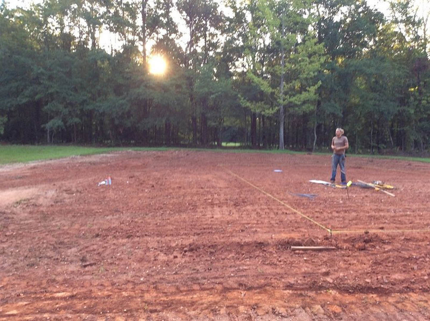 Dirt baseball field bordered by grass and trees, white chalk line visible, man standing near center under bright sunlight, clear blue sky overhead