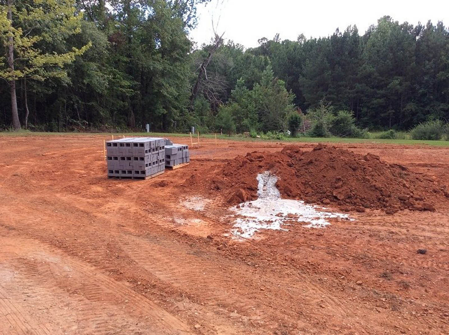 Muddy construction site with a pile of dirt, stacked grey concrete blocks, scattered cardboard boxes, and trees in the background