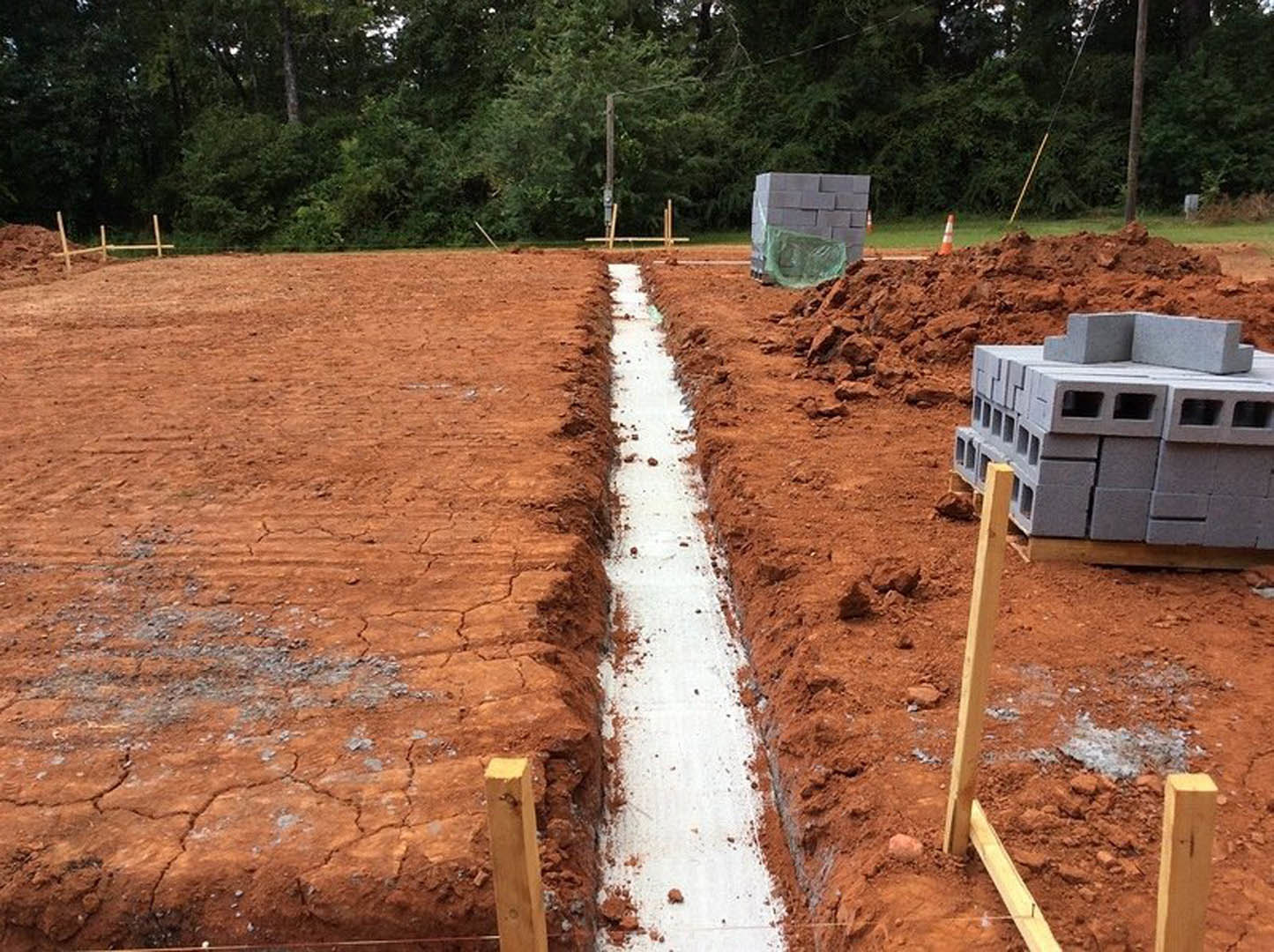 Dirt construction site with a deep trench, stacked grey concrete blocks, white gravel path, wooden post, and a pole-mounted light near trees
