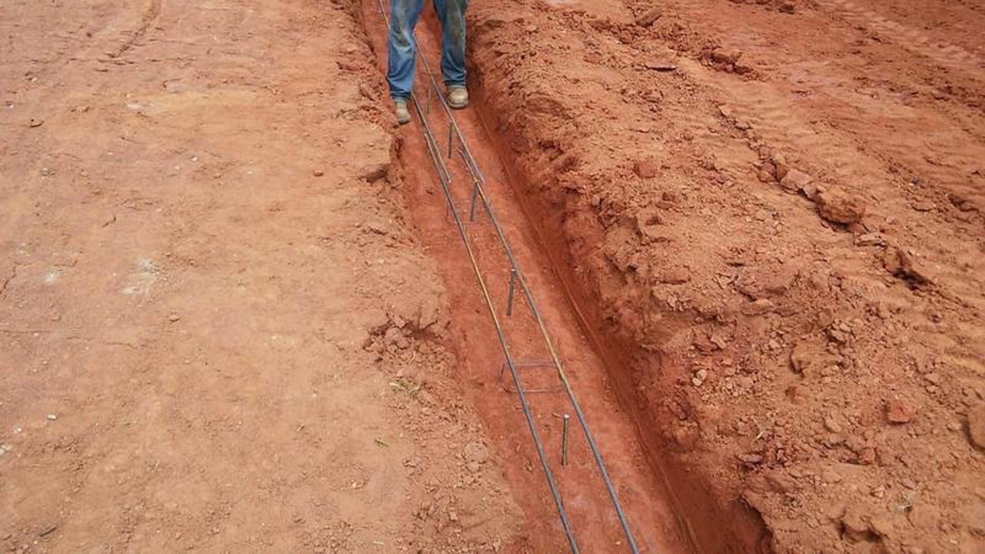 Man standing on a long metal rod in a dirt trench outdoors, soil and ground visible